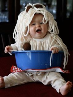 a baby sitting in a blue bowl with noodles on it's head and wearing a wig