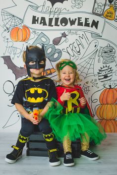 two children dressed up as batman and robin wayne sitting on a bench in front of a halloween themed wall