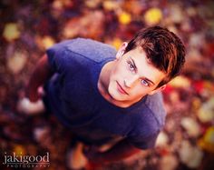 a young man looking up at the camera with blue eyes and brown hair, standing in front of leaves