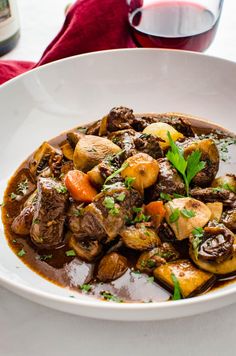 a white bowl filled with meat and vegetables on top of a table next to a glass of wine