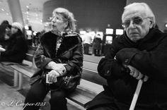 an old man and woman sitting on a bench in the middle of a busy airport