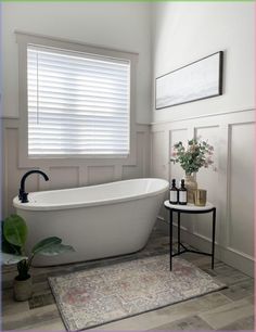 a white bath tub sitting under a window next to a table with a potted plant