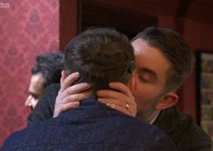 a man and woman kissing each other in front of a red wall with people looking on