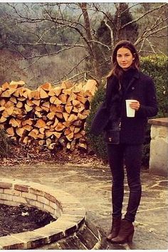 a woman standing next to a pile of firewood holding a coffee cup in her hand