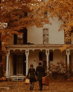 two people walking in front of a white house with autumn leaves on the ground and trees