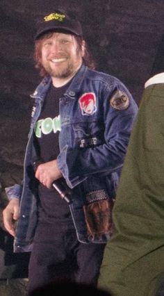 a man wearing a denim jacket and hat smiles while standing next to another man in a crowd