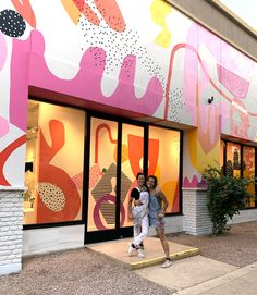 two women are standing in front of a store with colorful paintings on the side of it