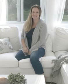 a woman sitting on top of a white couch in front of a window next to a coffee table