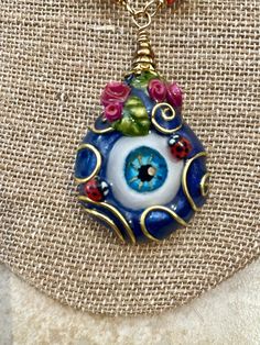 "Let the haters know you know their intentions with this over the top, more is more, evil eye nazar pendant. Even the two ladybugs standing guard beside the two tiny bouquets of fuscia roses are keeping watch over you. Large, dark blue pearlescent evil eye pendant is embellished with gold colored wire flourishes.  Every polymer clay evil eye pendant is sculpted and painted from scratch, starting with handcrafted, realistic-ish eye cabochons that have been marbled with dark blue, light blue, and Eye-catching Evil Eye Jewelry For Gift, Eye-catching Handmade Jewelry For Gifts, Unique Evil Eye Jewelry Gift, Unique Evil Eye Jewelry As A Gift, Unique Eye-shaped Jewelry For Gifts, Unique Evil Eye Jewelry For Festival, Unique Evil Eye Necklace For Gift, Handmade Spiritual Eye-shaped Jewelry, Nazar Amulet