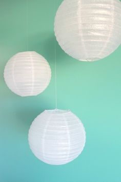 two white paper lanterns hanging from strings on a green and teal wall with blue background