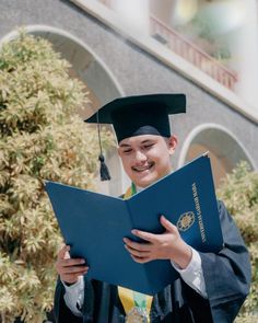 a young man in graduation gown holding a blue folder and smiling at the camera while wearing a cap and gown