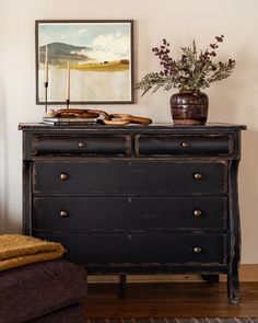 an old dresser with two drawers and a painting on the wall above it, along with a vase filled with flowers