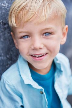 a young boy with blonde hair and blue eyes smiles at the camera while leaning against a wall