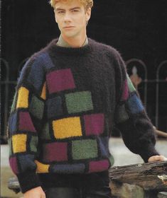 a young man wearing a multicolored sweater standing next to a tree trunk in front of a fence