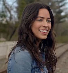 a woman with long dark hair and blue jean jacket smiles at the camera while standing on train tracks