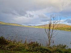 a body of water surrounded by trees and hills in the distance with clouds above it