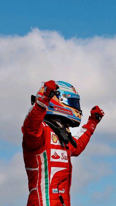 a man in a racing suit waves to the sky while holding his fist up and wearing a helmet