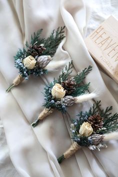 three white flowers and pine cones are placed on a bed sheet with a book in the background