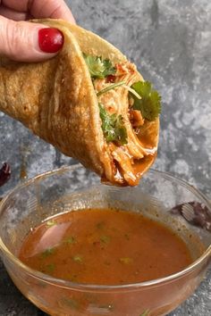 a hand holding a tortilla over a bowl of tomato soup and garnished with cilantro