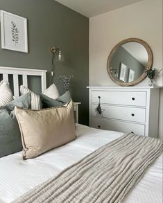 a white bed topped with pillows next to a dresser under a round mirror on top of a wall