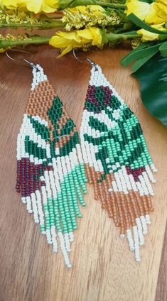 two green and brown beaded earrings sitting on top of a wooden table next to yellow flowers