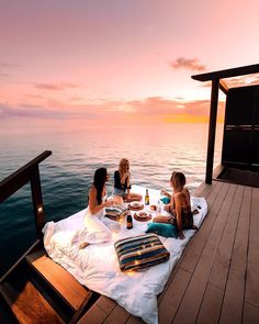 three women sitting on a dock at sunset eating food and drinking wine while the sun sets