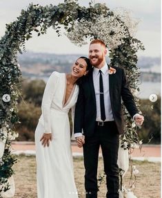 a newly married couple standing under an arch with greenery and white flowers on it