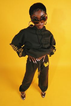 a young woman wearing sunglasses and a black hoodie stands in front of a yellow background