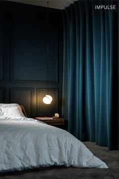 a bed sitting next to a window covered in blue drapes and a night stand with a lamp on it