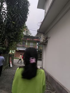 a woman in a green shirt walking down a street next to a white wall and trees