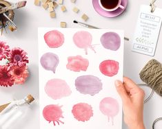 a person holding up a piece of paper with watercolor paint on it next to flowers and other crafting supplies