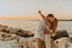 a man and woman standing on rocks near the water at sunset with their arms around each other