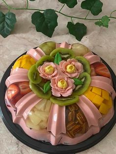 a cake decorated with fruit and flowers on top of a black plate next to green leaves