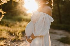 a pregnant woman in a white dress standing on a dirt road with the sun shining behind her