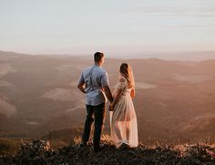 a man and woman standing on top of a mountain looking at each other while holding hands