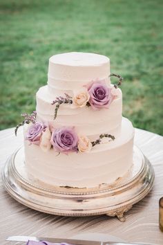 a three tiered cake with purple flowers on it sitting on top of a table