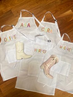 three white aprons with different names and shoes on them sitting on a wooden floor