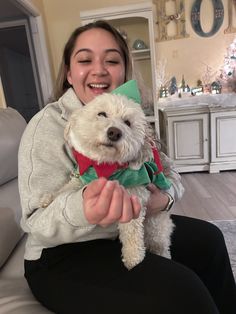 a woman sitting on a couch holding a white dog wearing a green bow around her neck