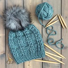 a knitted hat, yarn and knitting needles on a wooden surface with scissors next to it