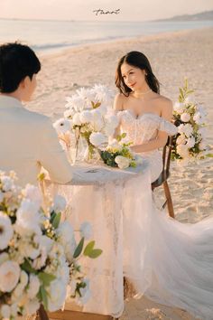 a man and woman sitting at a table on the beach with flowers in front of them