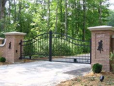 an iron gate and brick pillars in front of a driveway with trees on either side