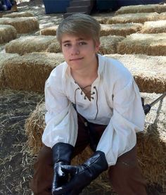 a young man sitting on top of hay bales wearing black gloves and a white shirt