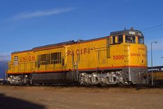 a yellow and red train traveling down tracks next to a dirt covered field on a sunny day