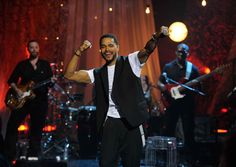 a man in black vest and white shirt on stage with his arms up while singing