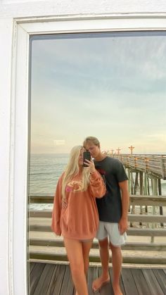 a man and woman standing on a pier taking a photo with their cell phone in front of them