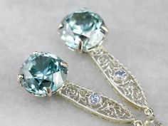 "Handcrafted in our New Hampshire studio, these earrings represent both something old, and something blue! The drops have been converted from an early 1900's era bar pin, set with light blue sapphires, and the filigree provides a pretty cobweb motif and gives length to the sparkling blue zircon gemstones we've set below. Metal: 14K White and Yellow Gold Gem: 2 Blue Zircon totaling 17.28 Carats Gem Measurements: 11 mm, Round Accents: 2 Sapphire totaling .14 Carats Earrings Length: 47 mm Earrings Heirloom Wedding Earrings Pierced, Vintage Drop Earrings With Prong Setting, Art Deco Gemstone Earrings For Gift, Heirloom Style Wedding Earrings With Intricate Design, Heirloom Wedding Earrings With Intricate Design, Blue Art Deco Wedding Earrings, Silver Gemstone Art Deco Earrings, Fine Jewelry Filigree Earrings For Wedding, Art Deco Silver Gemstone Earrings