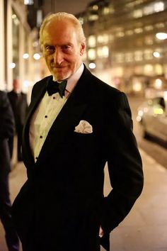 an older man in a tuxedo stands on the sidewalk at night with his hands in his pockets