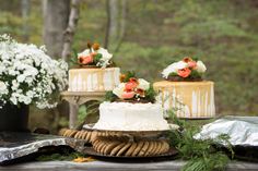 three wedding cakes with flowers on top sitting on a wooden table next to some cookies