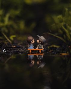 two small figurines sitting on a bench in the middle of a body of water