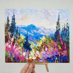 a painting being held up in front of a white wall with trees and mountains painted on it